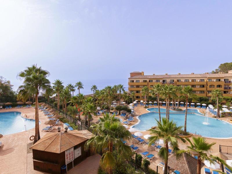 Großes Hotel mit mehreren Pools, Palmen und blauen Liegestühlen unter klarem Himmel.