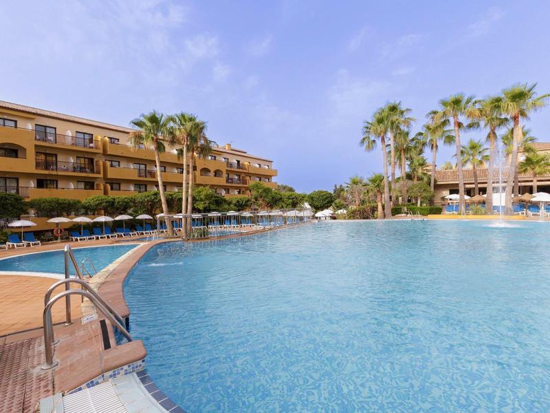 Großer Pool mit klarem Wasser, umgeben von Palmen und einem mehrstöckigen Hotelgebäude bei blauem Himmel.
