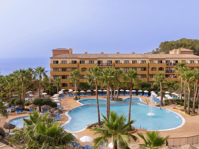 Luxushotel mit großem, geschwungenem Pool, Palmen und Meerblick im Hintergrund an einem klaren Tag.