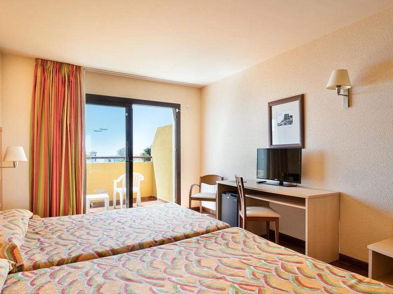 Hotelzimmer mit zwei Betten, Schreibtisch, Stuhl, Fernseher und Balkon mit Geländer bei Tageslicht.