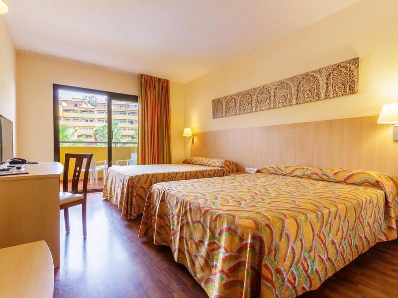 Hell eingerichtetes Hotelzimmer mit zwei Doppelbetten, Holzfußboden und Balkon mit Blick ins Grüne.