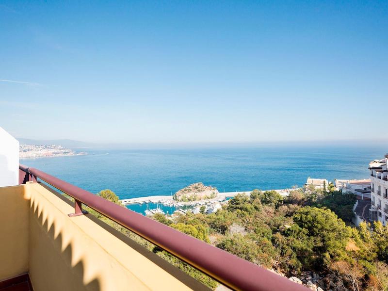 Blick von Balkon mit Geländer auf Meer, Küste, grüne Bäume und weiße Gebäude bei klarem Himmel.