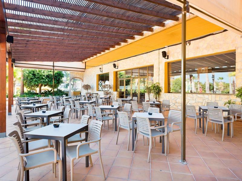 Freiluftrestaurant mit grauen Tischen und Stühlen, gelben Sonnensegeln und Terrakottaboden.