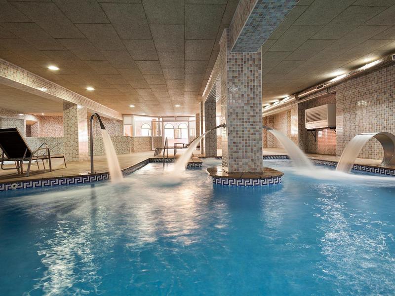 Innenraum mit großem Schwimmbecken, Wasserdüsen, Entspannungsbereich und warmem Licht.