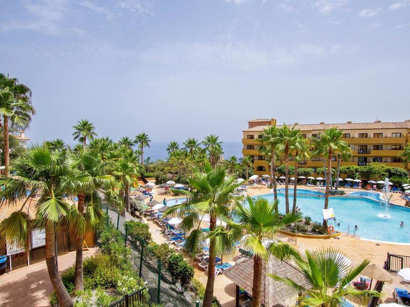 Großer Poolbereich mit Palmen, Liegestühlen und orangefarbenem Hotelgebäude im Hintergrund.