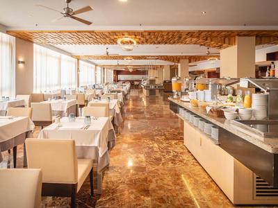 Großer, heller Speisesaal mit weißen Tischtüchern, beige Stühlen und Buffetbereich mit Saftspendern.