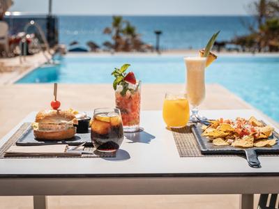 Tisch mit Getränken, Snacks und Cocktails am Pool mit Meerblick und Sonnenlicht.