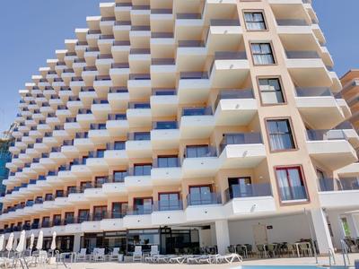 Modernes Hotel mit vielen Balkonen und großem, klar blauem Pool im Vordergrund bei sonnigem Himmel.