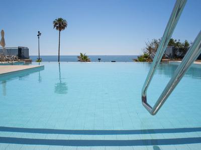 Klares Schwimmbad mit Edelstahlgeländer, Blick auf Meer und Palmen unter blauem Himmel.