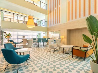 Moderner, heller Aufenthaltsraum mit bunten Sesseln, großen Fenstern und geometrischem Teppichmuster.