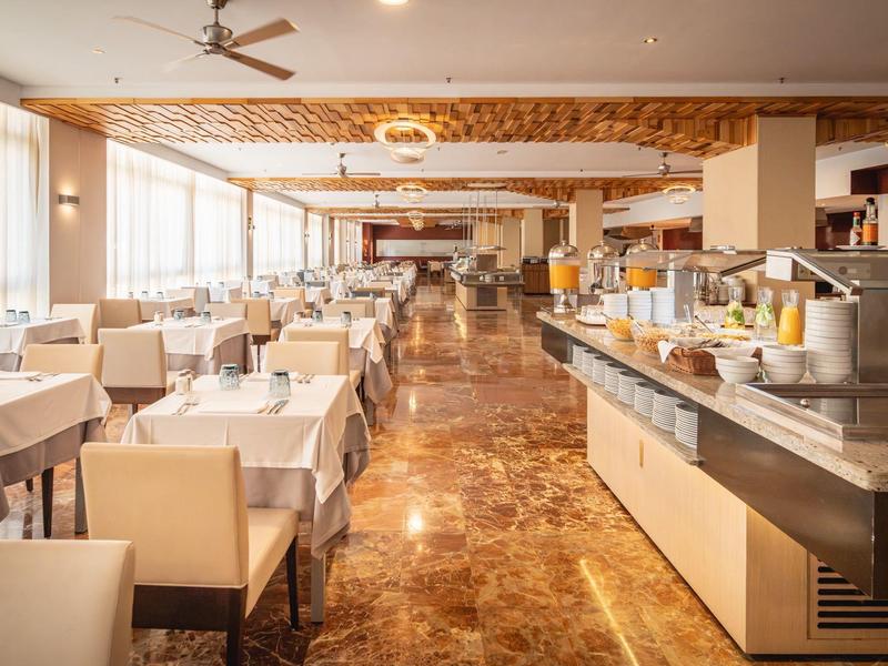 Großer, heller Speisesaal mit weißen Tischtüchern, beige Stühlen und Buffetbereich mit Saftspendern.