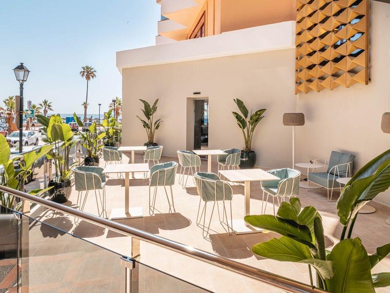 Helle Terrasse mit weißen Tischen und Stühlen, grünen Pflanzen und sonnigem Himmel im Hintergrund.
