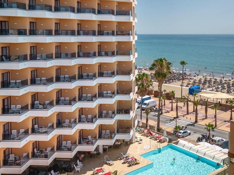 Hotel mit Balkonen, rechteckigem Pool, Palmen und Blick auf den Strand und das Meer.