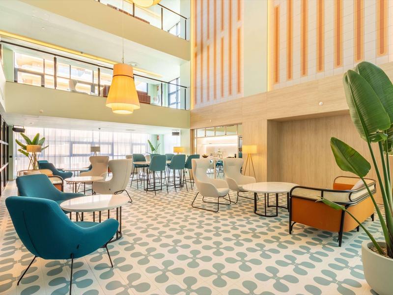 Moderner, heller Aufenthaltsraum mit bunten Sesseln, großen Fenstern und geometrischem Teppichmuster.