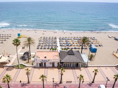 Strand mit blauer See, Sonnenschirmen, Liegen und Palmen entlang einer Promenade bei klarem Himmel.
