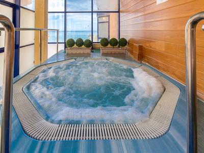 Innen-Whirlpool mit sprudelndem Wasser, blauer Umrandung und Holzwand, daneben Glasfenster und Pflanzen.