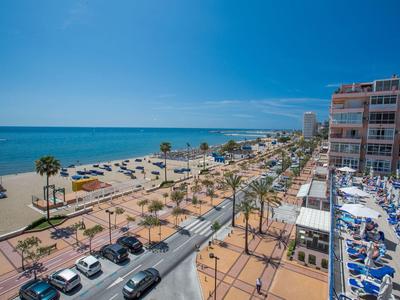 Strandpromenade mit Palmen, Parkplätzen, Meerblick und Hotels bei klarem Himmel.
