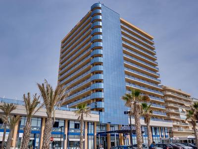 Modernes Hochhaus mit Glasfassade und umlaufenden Balkonen, umgeben von Palmen und klarem Himmel.