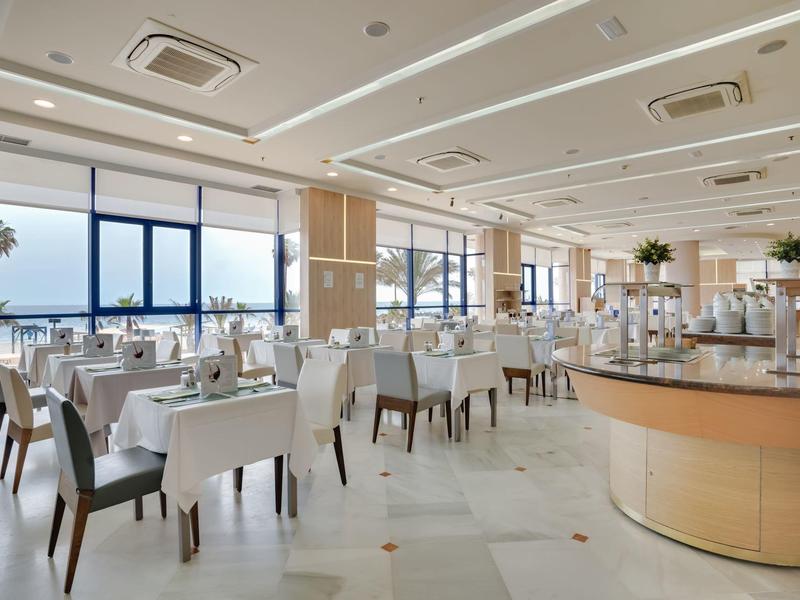 Großer, heller Speisesaal mit weißen Tischen, Holzstühlen und Panoramafenstern mit Meerblick.