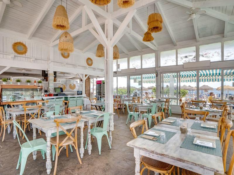 Heller, luftiger Gastraum mit weißen Holzbalken, Pastelltischen und Stühlen, großen Fenstern.