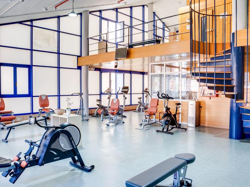 Großer heller Fitnessraum mit verschiedenen Trainingsgeräten und blauen Metalltreppen.