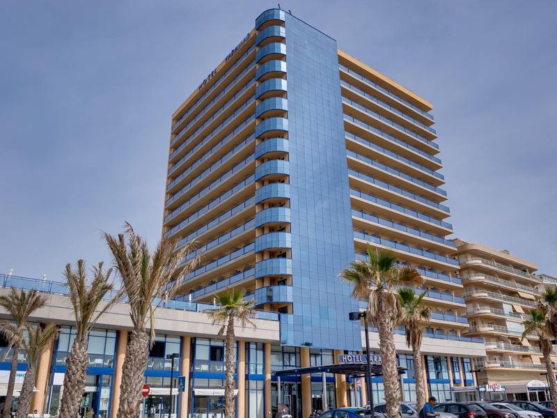 Modernes Hochhaus mit Glasfassade und umlaufenden Balkonen, umgeben von Palmen und klarem Himmel.