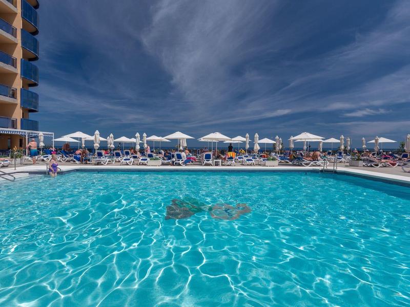Großer Außenpool mit klarem blauem Wasser, Sonnenschirmen und Liegestühlen unter bewölktem Himmel.
