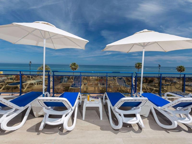 Mehrere weiße Liegestühle mit blauen Auflagen unter weißen Sonnenschirmen vor Meer und blauem Himmel.