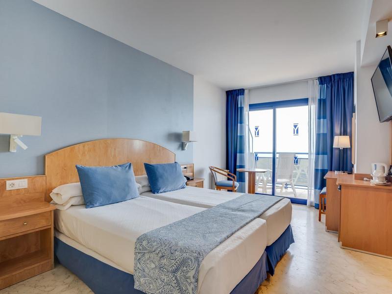 Hell eingerichtetes Hotelzimmer mit Doppelbett, blauem Farbakzent und Balkon mit Meerblick.