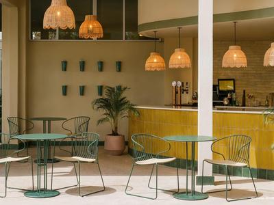 Area lounge di hotel moderna con bancone giallo, sedie verdi e lampade sospese in un ambiente luminoso.