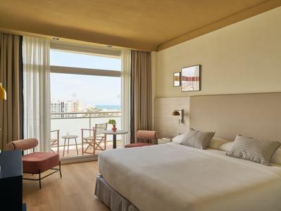 Chambre d'hôtel lumineuse avec lit double, chaises et balcon avec vue sur la mer.