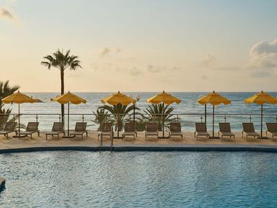 Piscine avec eau bleue, entourée de chaises longues avec parasols jaunes et palmiers au bord de la mer
