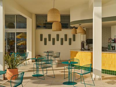 Terrasse d'hôtel lumineuse avec murs jaunes, chaises et tables vertes, lampes naturelles et plantes.