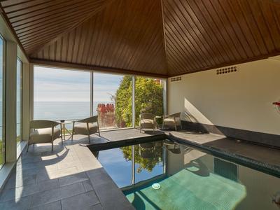 Piscine intérieure avec grandes fenêtres donnant sur la mer et le jardin, plafond en bois, ambiance lumineuse.