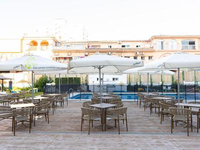 Terrazza con sedie e tavoli, grande piscina e ombrelloni davanti a un edificio alberghiero.