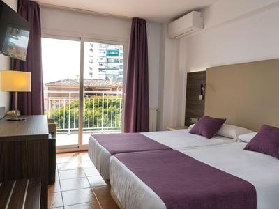Nowoczesny pokój hotelowy z podwójnym łóżkiem, fioletowymi akcentami i balkonem z widokiem na miejskie budynki.