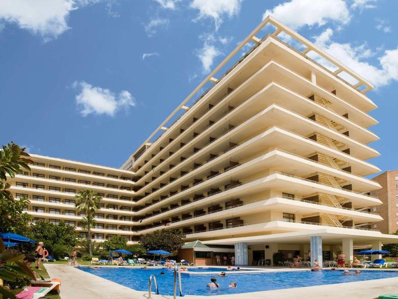 Großes, helles Hotel mit Balkonfronten und blauem Pool unter blauem Himmel.