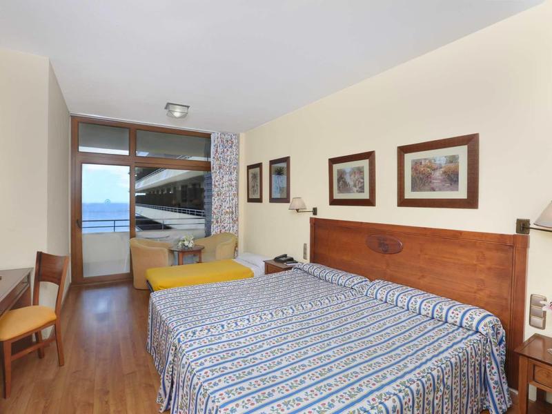 Hell eingerichtetes Schlafzimmer mit Doppelbett, Holzmöbeln und Blick auf das Meer durch Fenster.