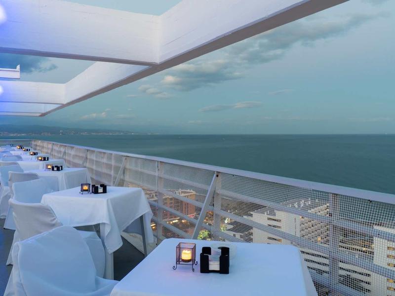 Balkon mit Tischen und weißen Stühlen, Blick auf das Meer und bewölkten Himmel bei Dämmerung.