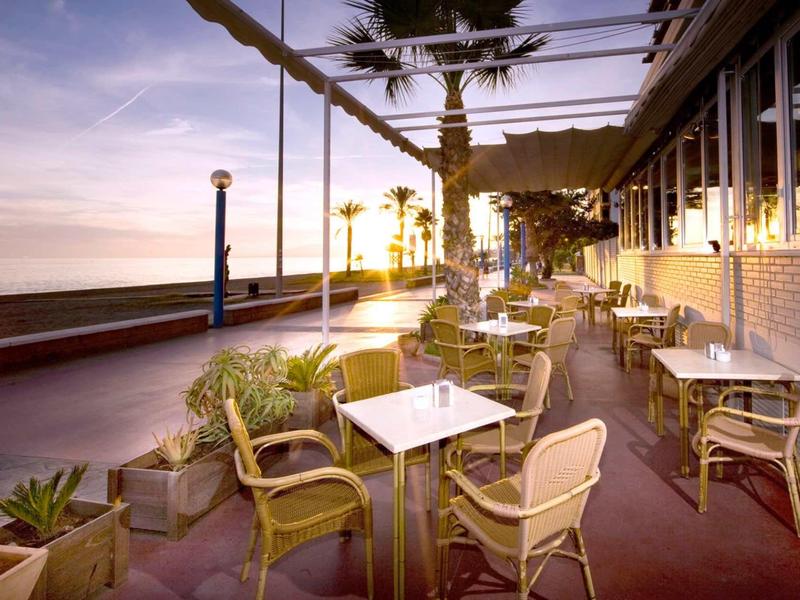 Terrassen-Café mit Tischen und Stühlen, Palmen und Meer im Sonnenuntergang im Hintergrund.