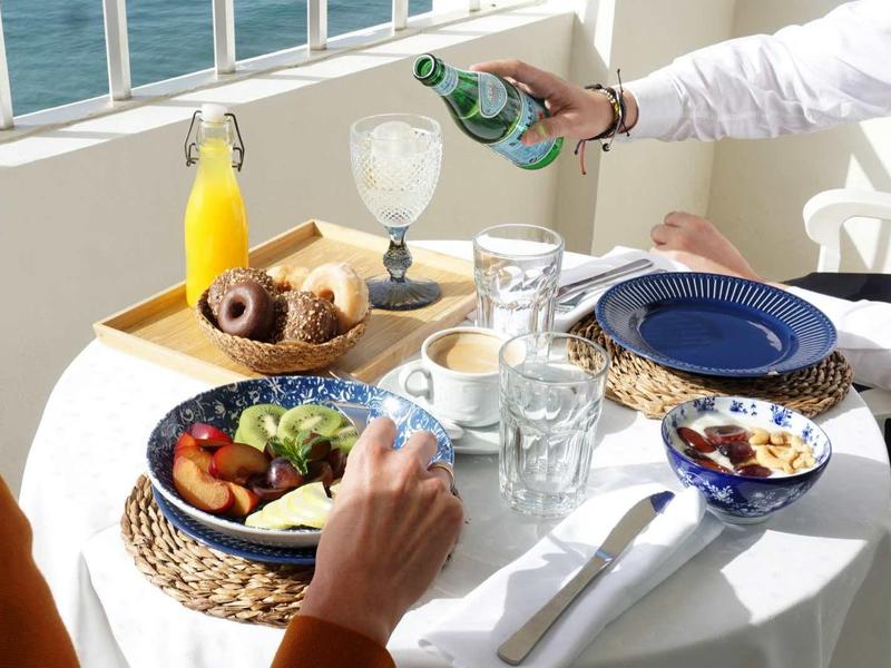 Zwei Personen genießen Frühstück mit Obst, Müsli und Getränken an einem sonnigen Tisch am Meer.
