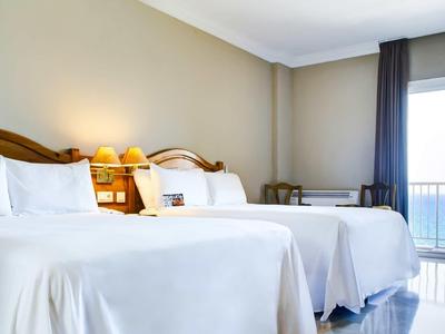 Helles Hotelzimmer mit Doppelbetten, Holzkopfteil und Blick aufs Meer durch große Fenster.