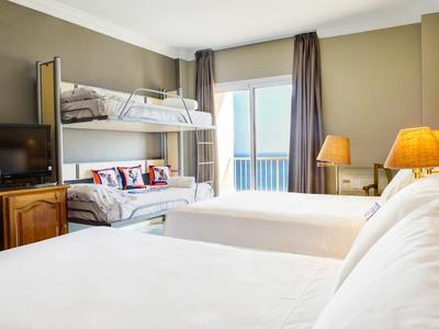 Helles Hotelzimmer mit Doppelbett, Etagenbett, Fernseher und Balkon mit Vorhängen.