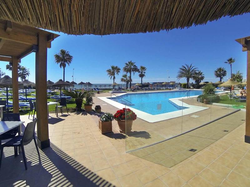 Schwimmbad mit klar blauem Wasser, Sonnendach, Stühlen, Palmen und klarem Himmel.