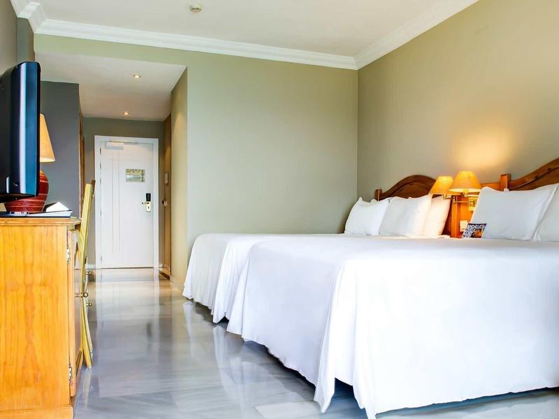 Hotelzimmer mit zwei Doppelbetten, Holzmöbeln, Flachbildfernseher und hellen Wänden.