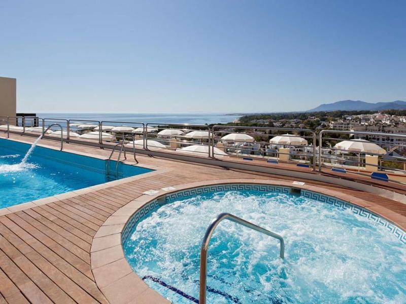 Rundlicher Whirlpool und rechteckiger Pool mit Holzdeck und Meerblick unter klarem Himmel.