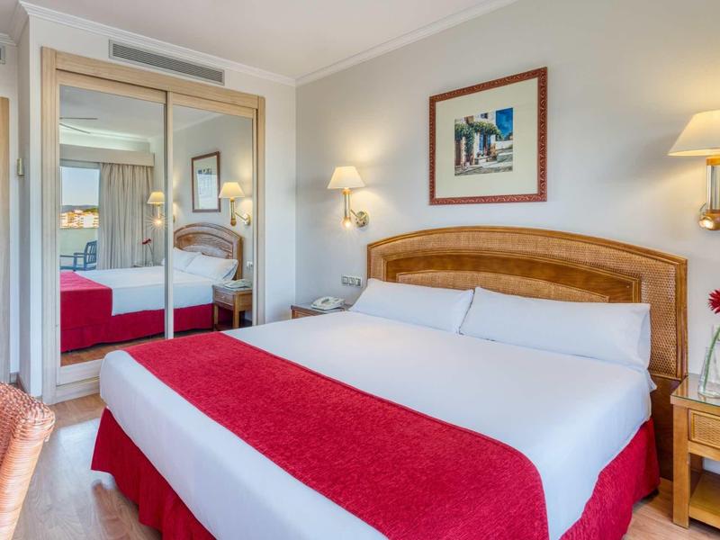 Helles Hotelzimmer mit großem Bett, weißen und roten Bettenbezügen, Holzmöbeln und Spiegelschrank.