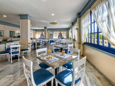 Bright restaurant with white furniture, blue chairs, and large windows with yellow curtains.