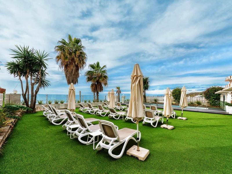 Grüner Rasen mit weißen Liegestühlen und Sonnenschirmen vor blauem Himmel und Palmen.