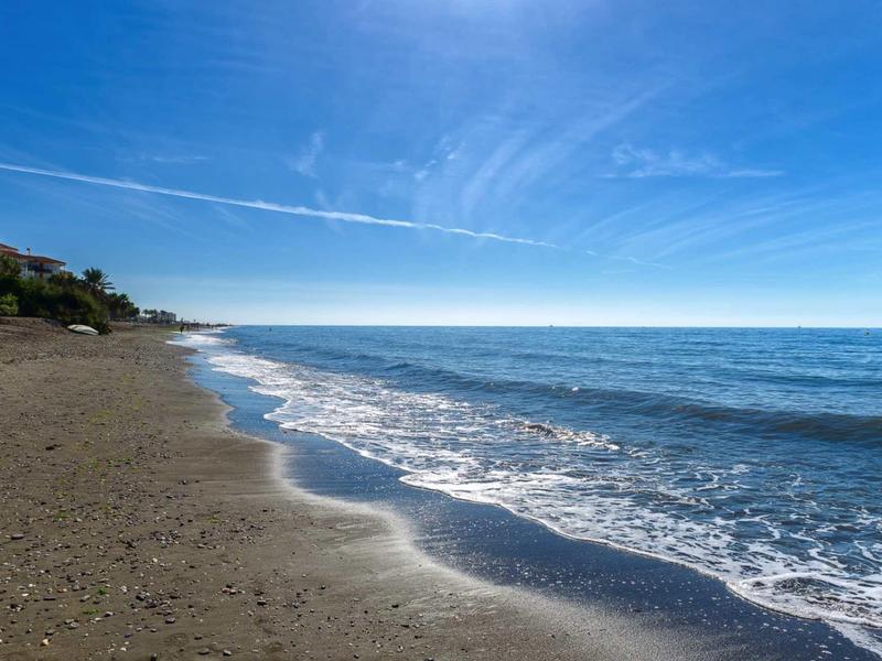 Strand mit sanften Wellen, blauem Himmel und wenigen Häusern im Hintergrund am linken Rand.